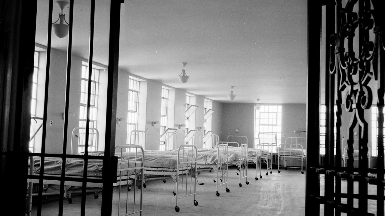 A black-and-white photo from 1938 of a large hospital ward room in which about ten narrow wheeled hospital beds are lined up before a windowed wall. A heavy wrought-iron gate, like old-fashioned prison bars and presumably used to lock patients in, has been opened to allow the photo to be taken.