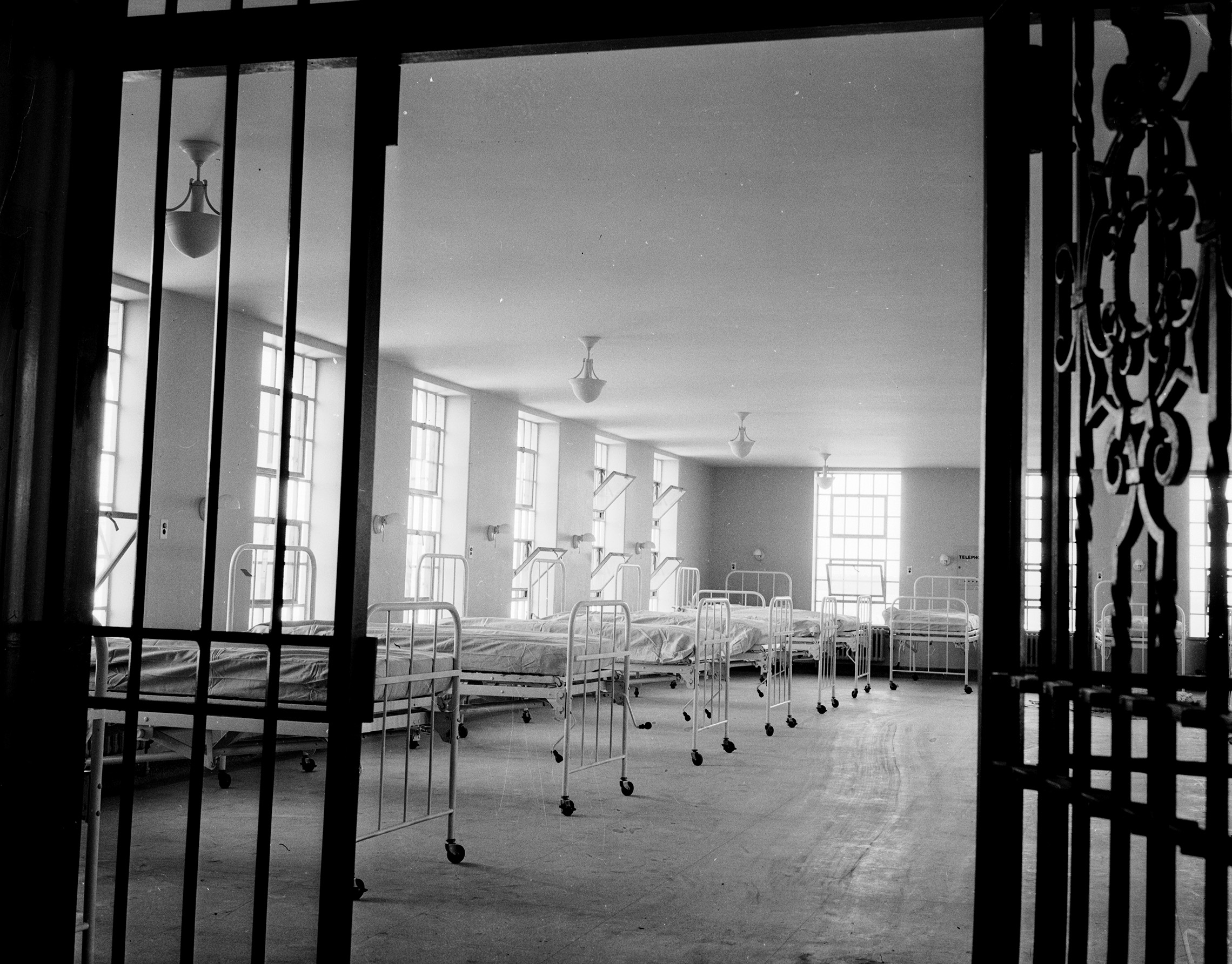 A black-and-white photo from 1938 of a large hospital ward room in which about ten narrow wheeled hospital beds are lined up before a windowed wall. A heavy wrought-iron gate, like old-fashioned prison bars and presumably used to lock patients in, has been opened to allow the photo to be taken.